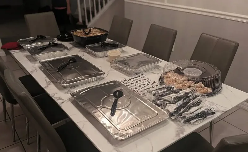 Covered catering trays, serving utensils, and sides arranged across a dining table